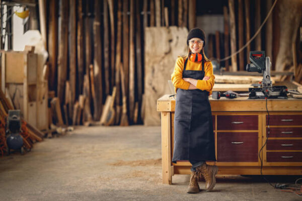 Full body of smiling confident young female joiner in apron standing near workbench and looking at camera friendly while working in craft workshop