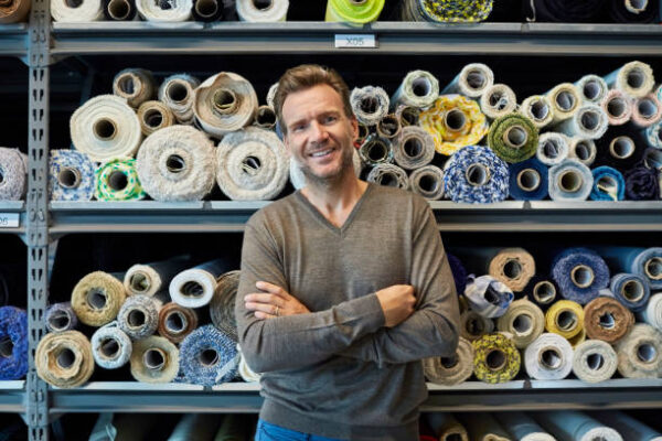 Portrait of confident businessman with arms crossed against rolled fabric. Executive is smiling while standing against shelves. He is wearing smart casuals in textile factory.